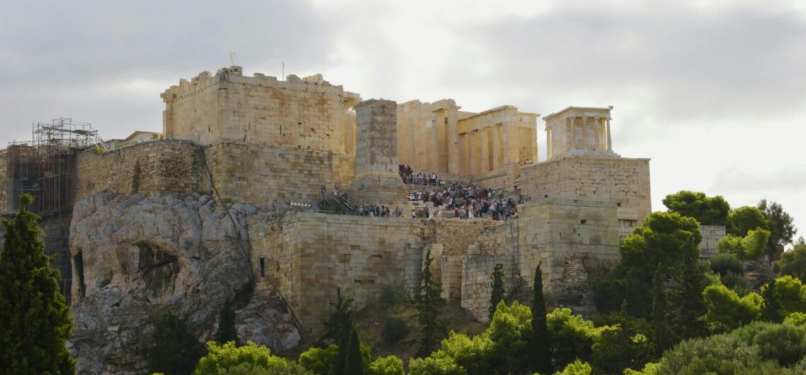 Ancient Acropolis ruins atop a rocky hill with visitors climbing the limestone steps, surrounded by trees beneath a cloudy sky.