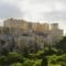 Ancient Acropolis ruins atop a rocky hill with visitors climbing the limestone steps, surrounded by trees beneath a cloudy sky.