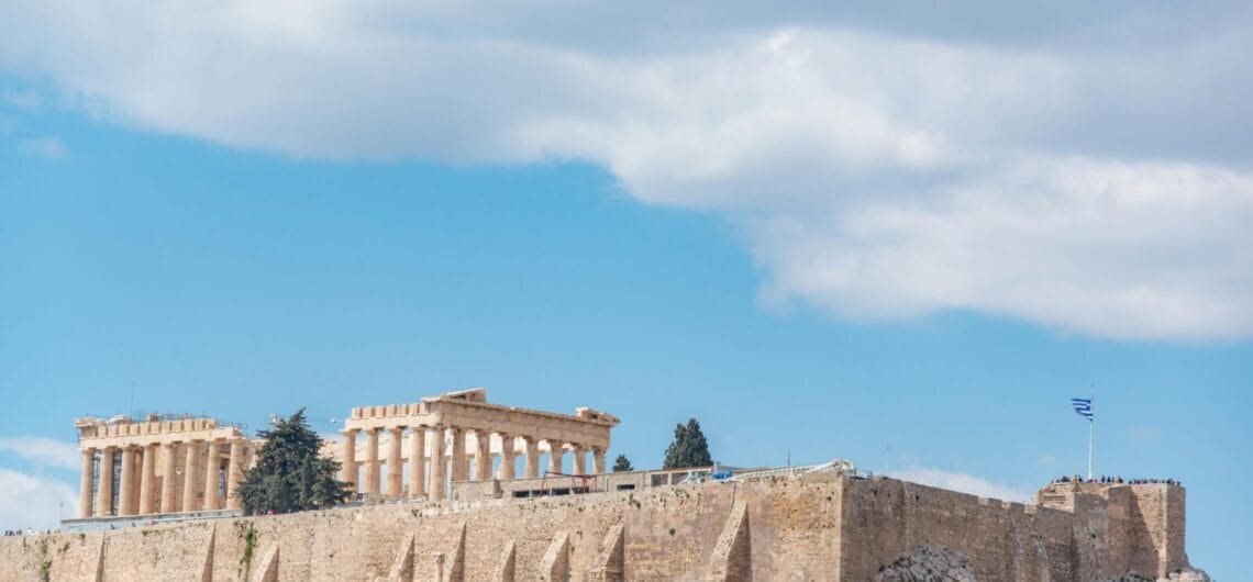 Ancient Parthenon temple atop the Acropolis with a stone fortress wall and a Greek flag against a blue sky for context.
