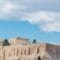 Ancient Parthenon temple atop the Acropolis with a stone fortress wall and a Greek flag against a blue sky for context.