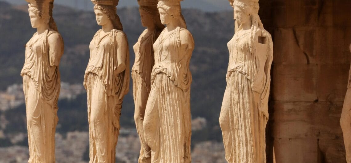 Row of carved female figures (caryatids) acting as columns along a stone frieze, with a distant landscape behind them in warm sunlight.