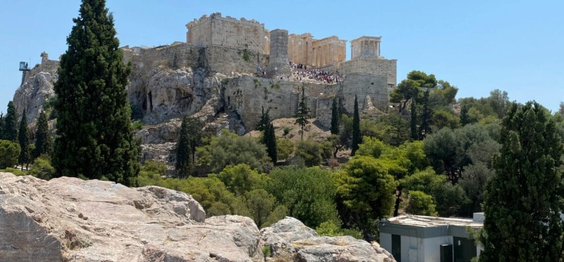 Ancient Parthenon ruins atop a rocky hill with surrounding trees under a clear blue sky.