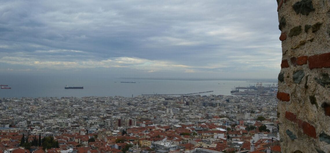 Coastal city viewed from above with dense red-tiled roofs and ships on the calm bay; a stone wall on the right frames the scene.