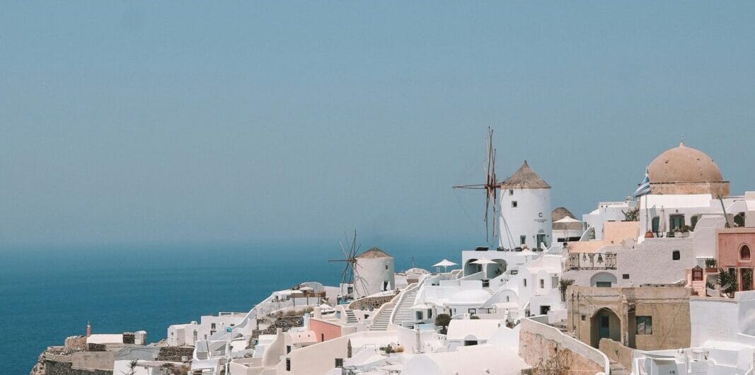 White-washed hillside village with windmills and blue domes overlooking the blue Aegean Sea.