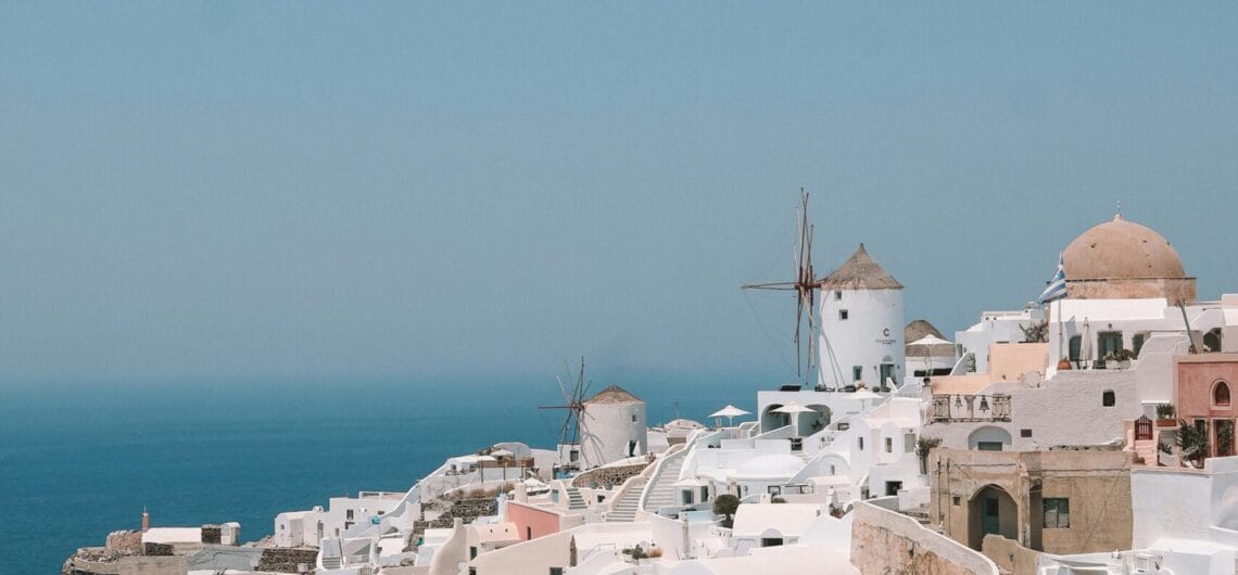 Whitewashed Cycladic buildings with domed roofs cascading down a cliff toward the blue sea and sky, Santorini, Greece.