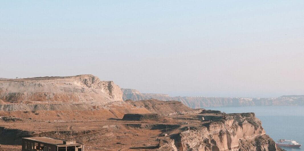 Abandoned concrete building perched on a rocky coastal cliff with dry scrub and a distant sea horizon.