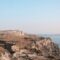 Abandoned concrete building perched on a rocky coastal cliff with dry scrub and a distant sea horizon.