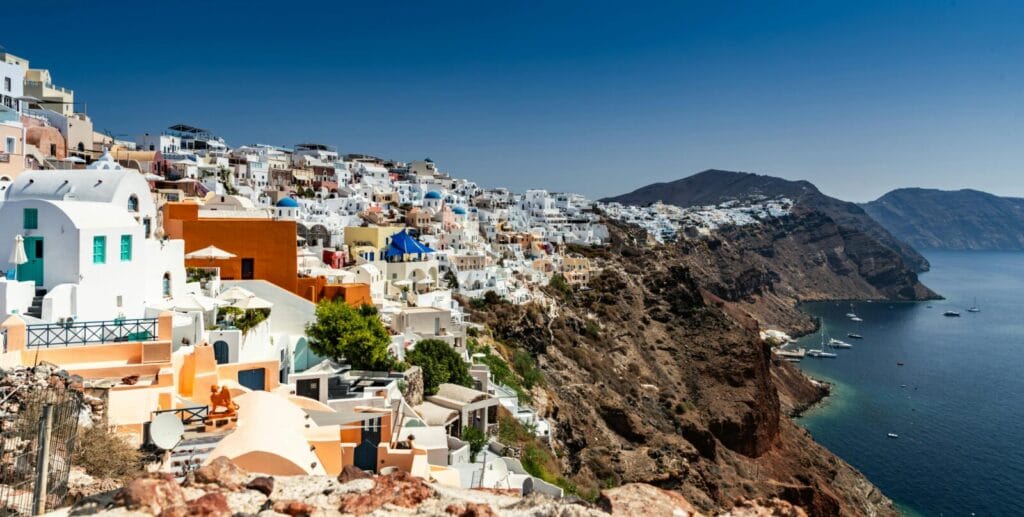 White-washed houses and blue-domed churches cling to a cliff above the deep blue Aegean Sea in Santorini.](https://)