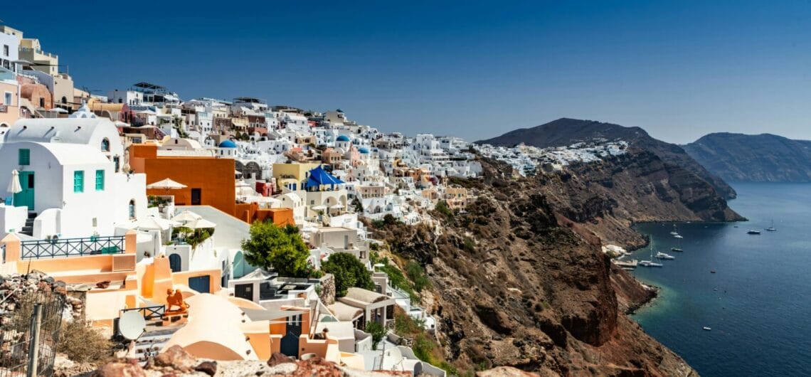 White-washed houses and blue-domed churches cling to a cliff above the deep blue Aegean Sea in Santorini.](https://)