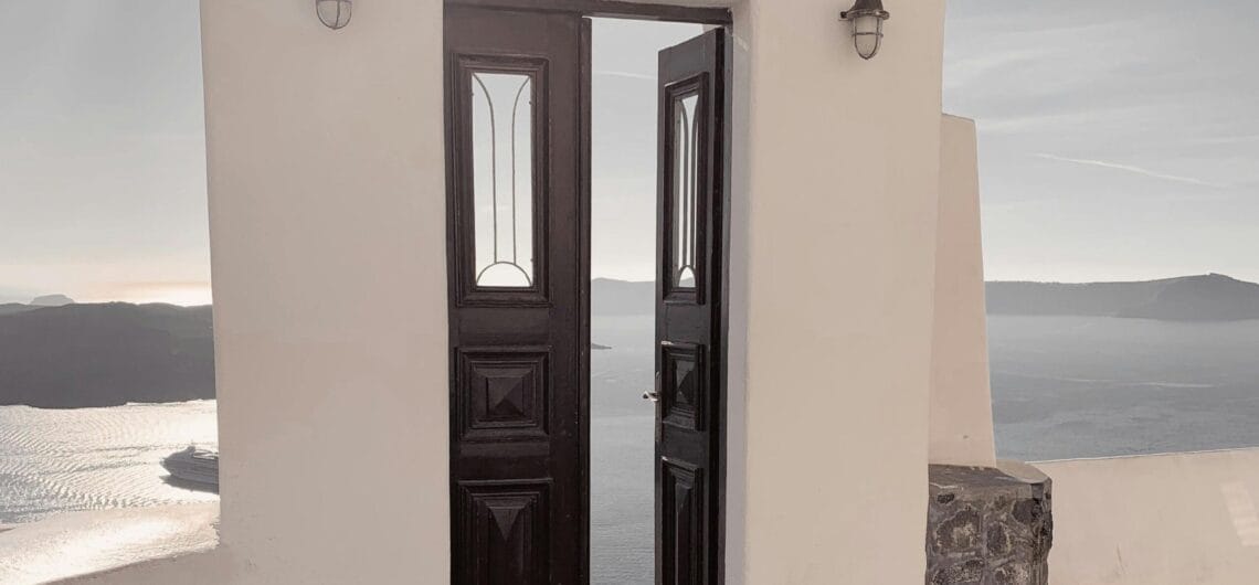White stucco wall with a dark double door opening to a sea view; two lantern-style lights flank the doorway against a bright sky. Alternative: An arched doorway set in a white wall overlooking the water, with two outdoor lamps on either side.