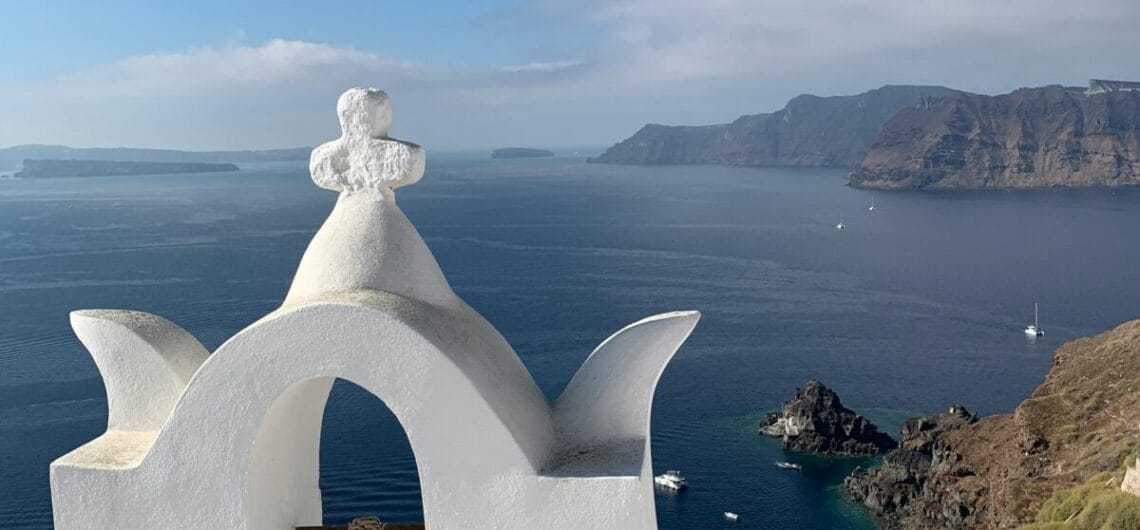 White bell tower with a small cross on top, overlooking blue Aegean Sea and rugged cliffs of Santorini.