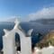White bell tower with a small cross on top, overlooking blue Aegean Sea and rugged cliffs of Santorini.