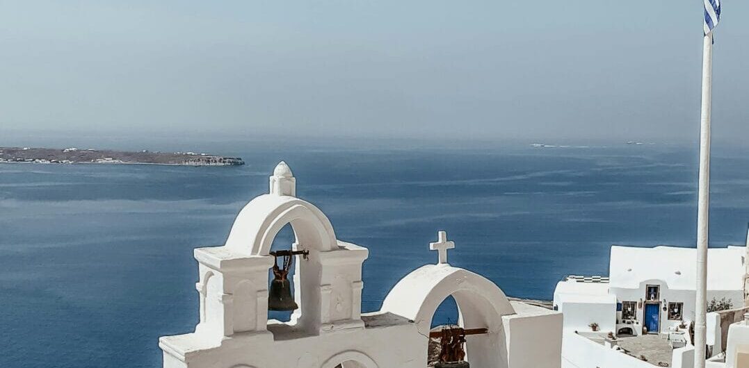 Whitewashed bell arches on a cliff overlooking the blue Aegean Sea, with a Greek flag nearby.