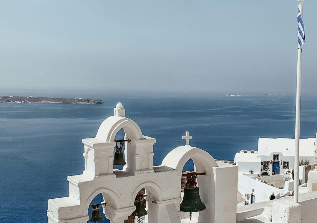 Whitewashed bell arches on a cliff overlooking the blue Aegean Sea, with a Greek flag nearby.