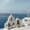 Whitewashed bell arches on a cliff overlooking the blue Aegean Sea, with a Greek flag nearby.