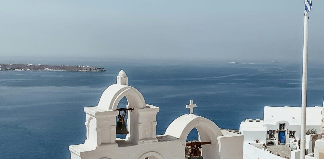 Whitewashed church bells in a Greek island setting with the Aegean Sea in the background and a Greek flag on a tall pole nearby