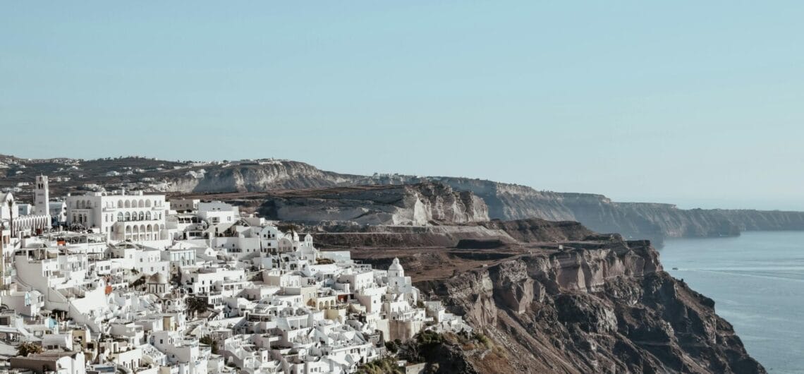 White-washed buildings cling to a cliffside town overlooking the blue sea and distant cliffs.